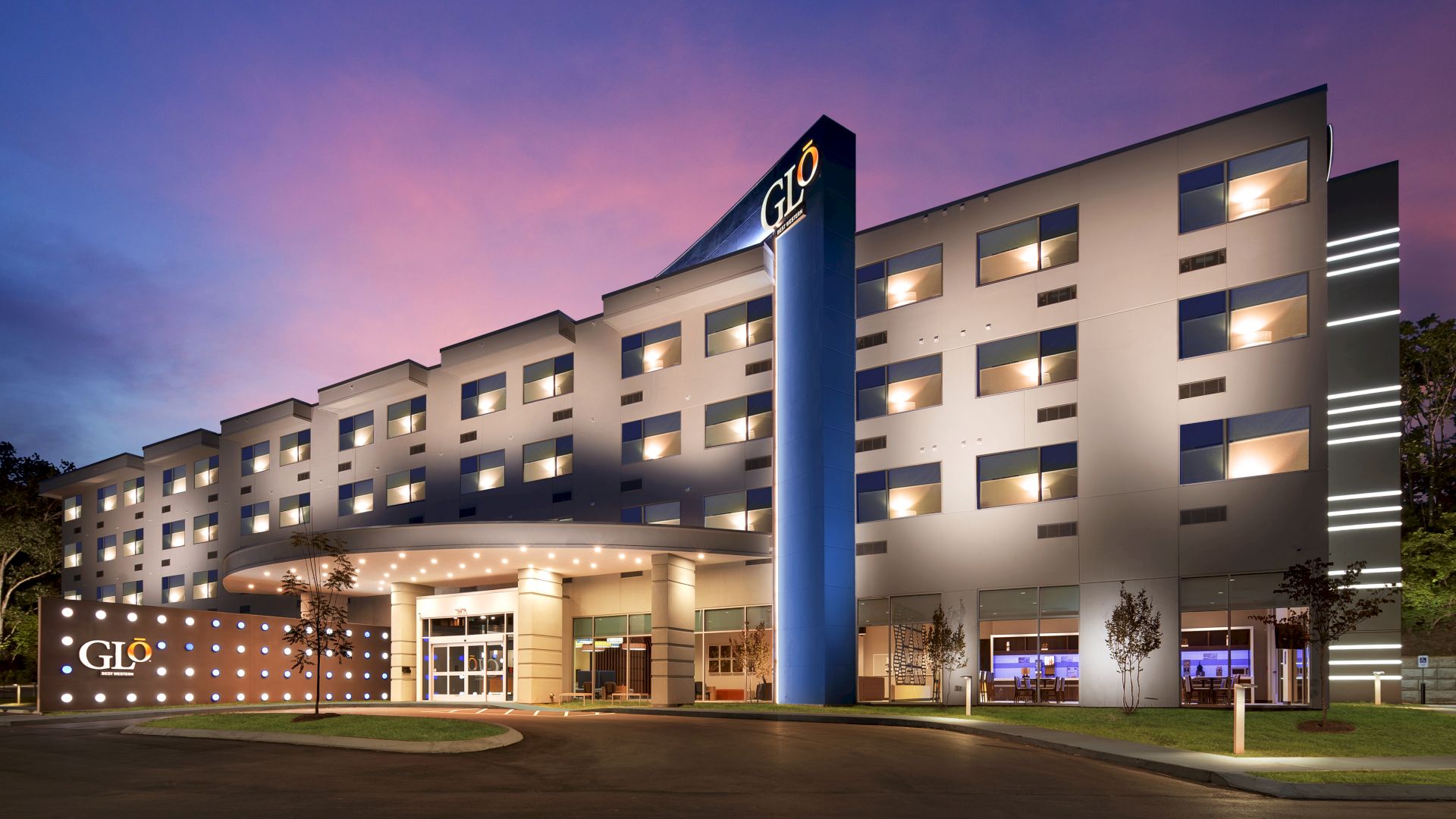 The image shows a modern hotel building at dusk with the sign "Glo" prominently displayed, featuring a well-lit facade and entrance.