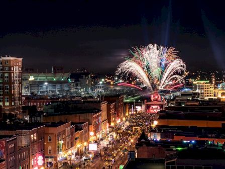 The image shows a vibrant cityscape at night with buildings and streets illuminated, and colorful fireworks lighting up the sky above.