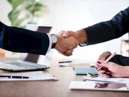 Two individuals in formal attire are shaking hands over a desk, with paperwork, a laptop, and a pen in the scene.