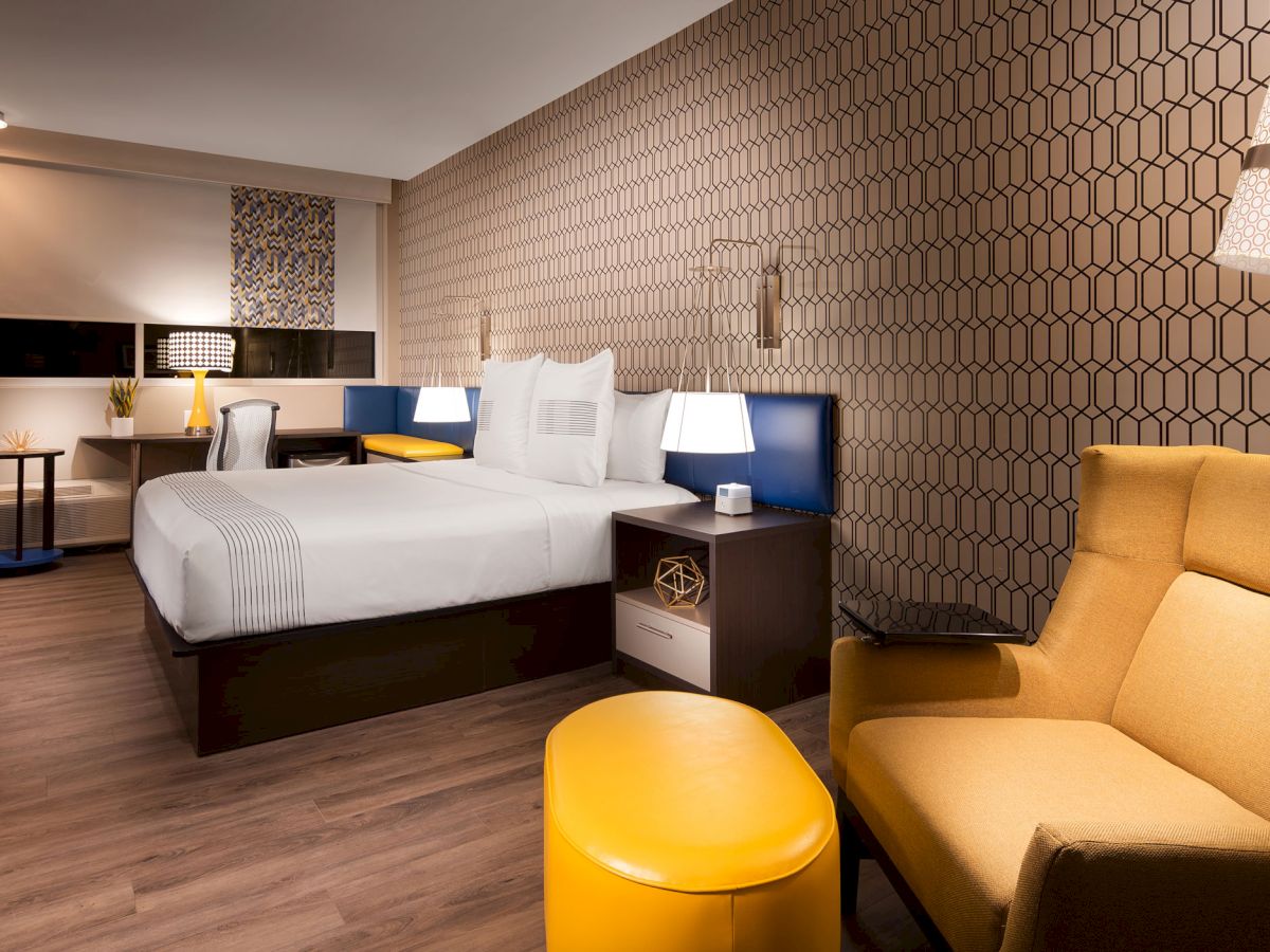 A modern hotel room features a bed, chairs, a table, lamps, a TV, and patterned wallpaper, with a cozy and stylish interior design.