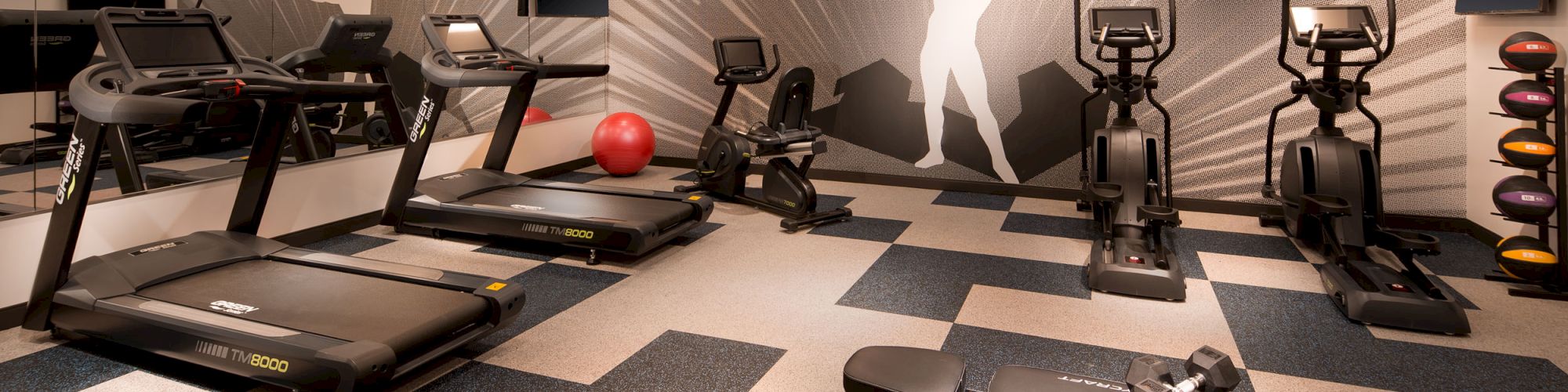 This image shows a modern gym with treadmills, stationary bikes, a weight bench, dumbbells, and a large wall mural of a weightlifter at the back.