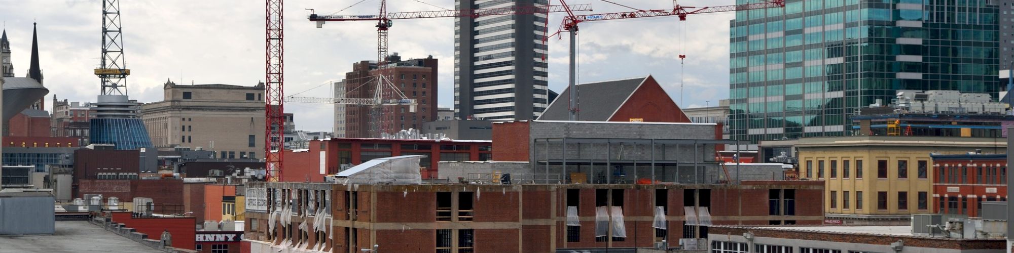 The image shows an urban cityscape with various buildings, construction cranes, and a mix of older brick structures and modern high-rises, under a cloudy sky.