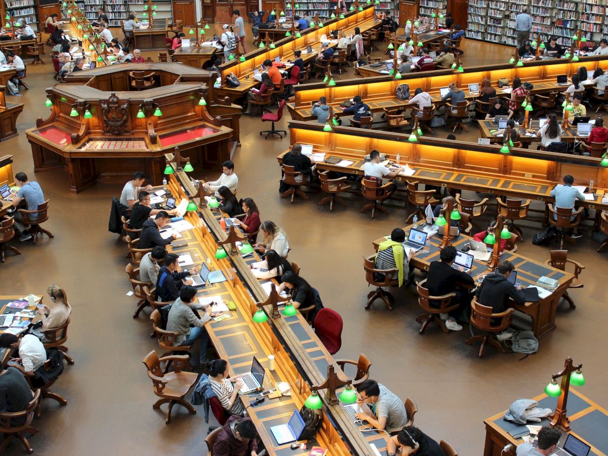 A large library room filled with numerous people studying or reading at long tables, with green desk lamps and bookshelves lining the walls.