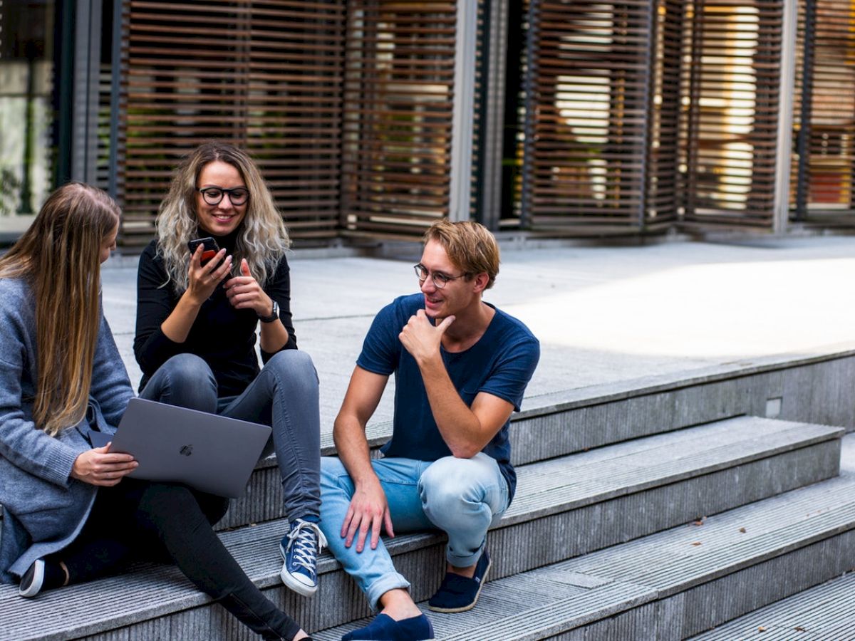 Three people are sitting on outdoor steps, engaging with a laptop and a phone, while casually talking and smiling.