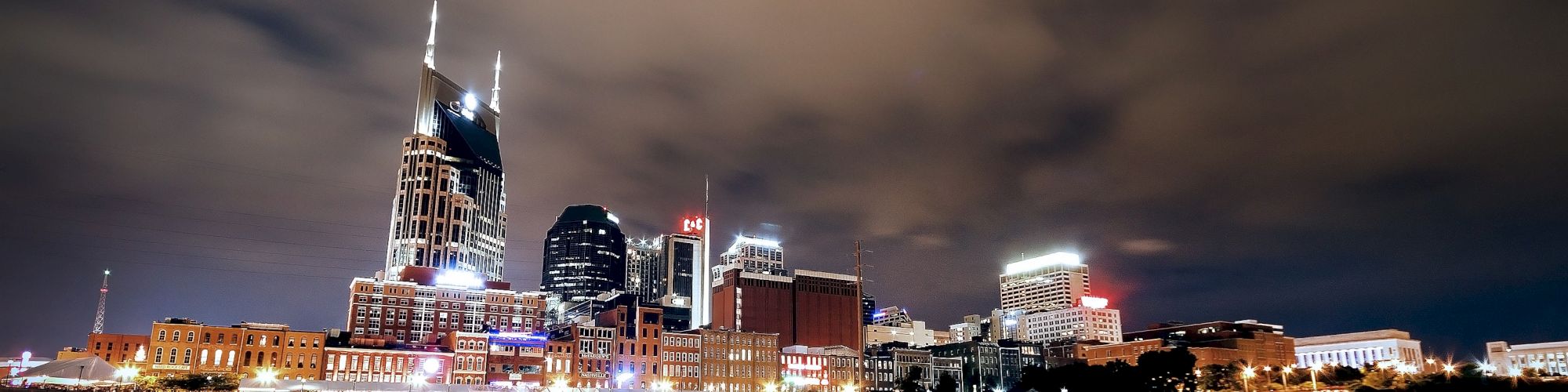 The image showcases a nighttime city skyline with illuminated buildings reflecting in the water below, under a cloudy sky.