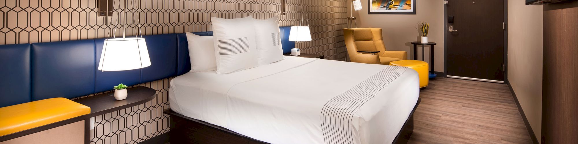 A modern hotel room with a double bed, yellow armchair, small side tables, lamps, and geometric-patterned wallpaper, featuring wooden floors.