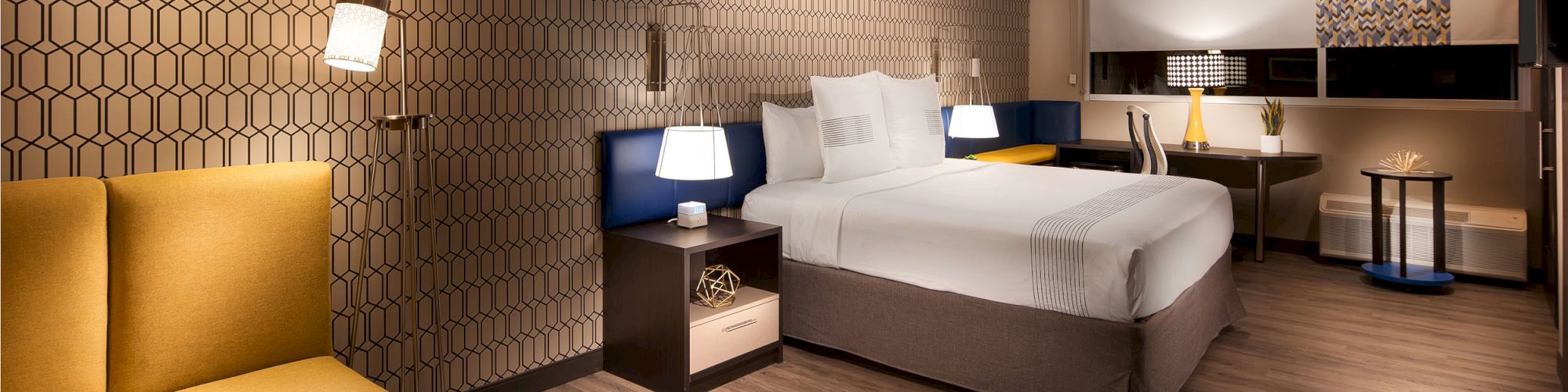 A modern hotel room features a bed, nightstands, lamps, a seating area, and a desk with a chair. The decor includes patterned wallpaper and wooden flooring.