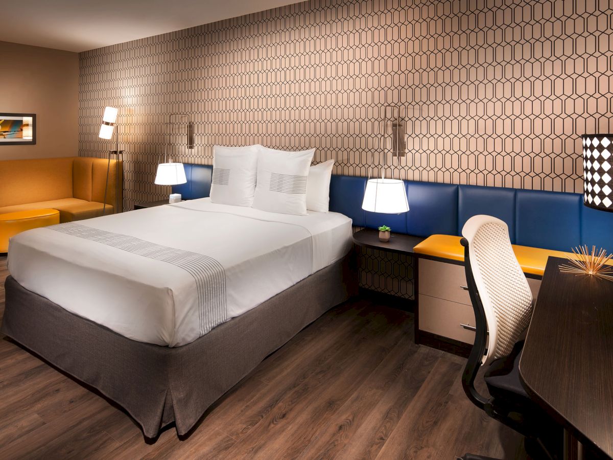 A modern hotel room features a bed, nightstands, a desk, a chair, and a cozy seating area, all with geometric decor accents and vibrant lighting.