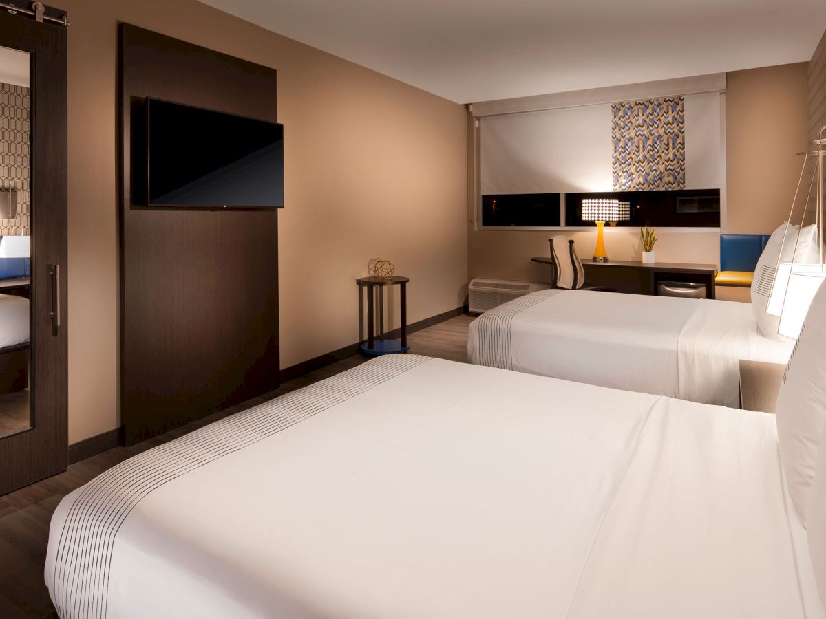 The image shows a modern hotel room with two double beds, a wall-mounted TV, a work desk with a lamp, and contemporary decor.