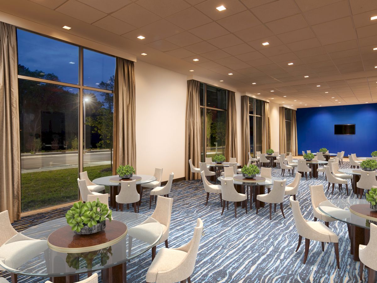 A modern, well-lit room with large windows, featuring multiple round tables with white chairs and small green plants as centerpieces, against a blue accent wall.