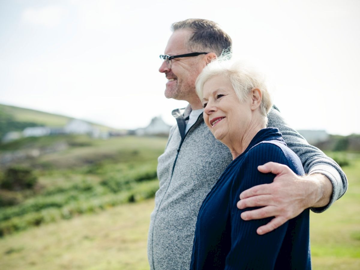 An older couple is standing outdoors, smiling and embracing, with a grassy hill and a few buildings in the background.