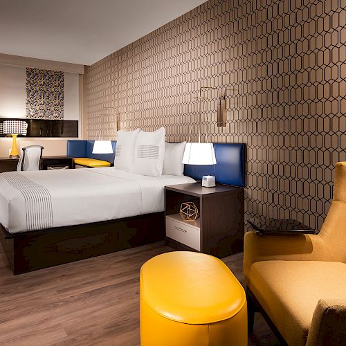 This image shows a modern hotel room with a bed, yellow armchair, matching ottoman, desk, and lamps, featuring geometric wallpaper.