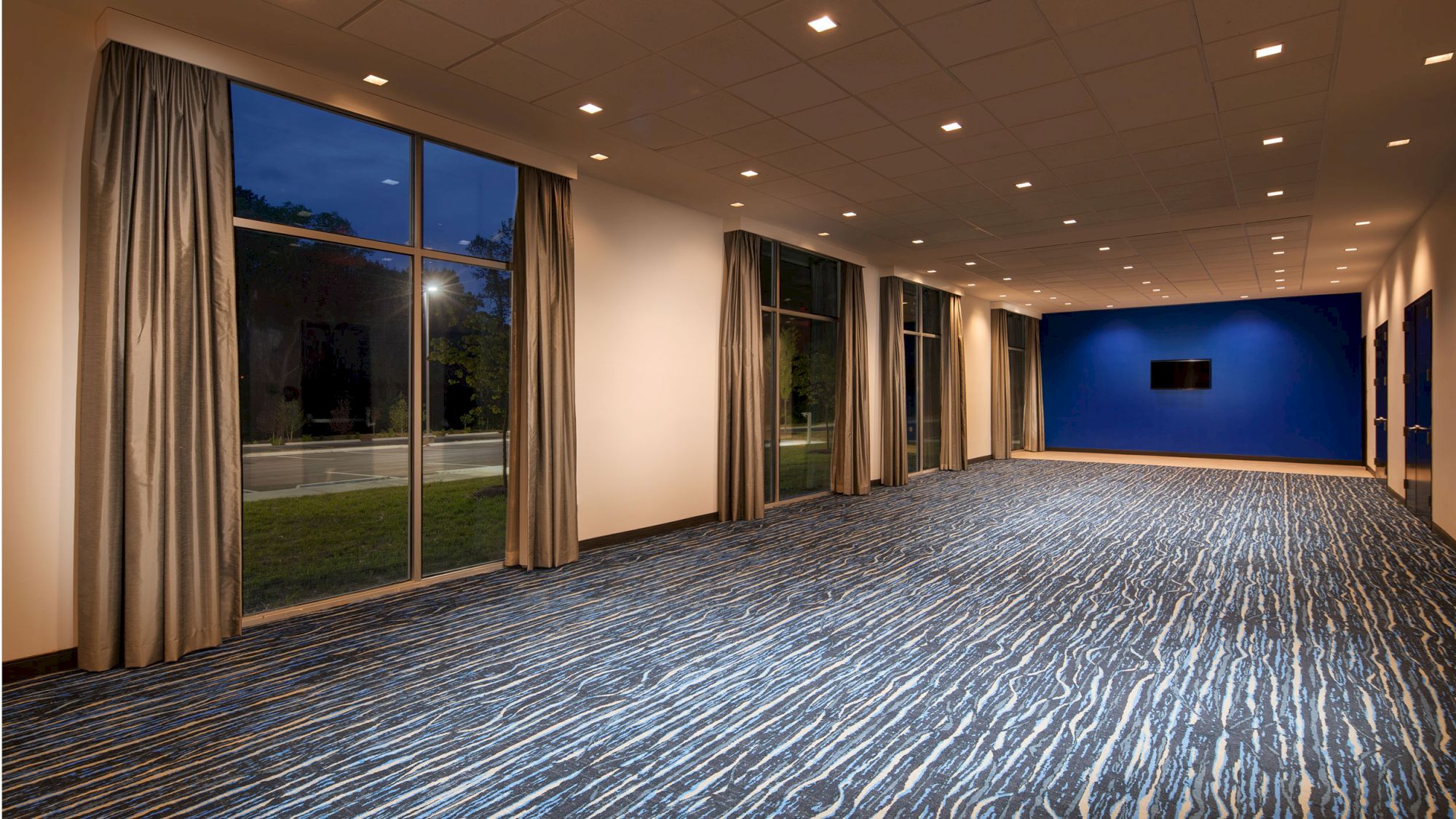 A spacious conference room with large windows, blue accent wall, ceiling lights, patterned carpet, and curtains overlooking a grassy outdoor area.