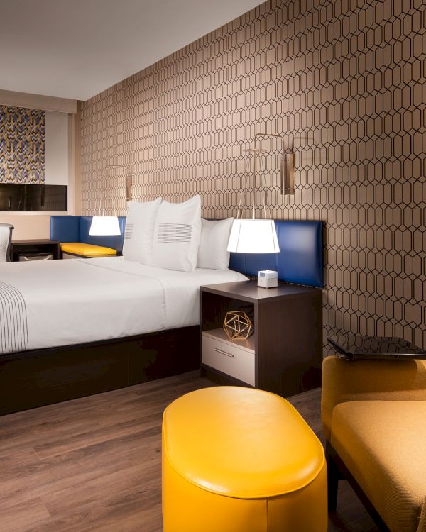 A modern hotel room with a bed, yellow chair, and geometric wallpaper, featuring ambient lighting and a cozy atmosphere.