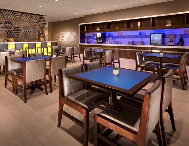 The image shows a modern dining area with tables, chairs, and a bar. The lighting is warm, and the decor is contemporary and clean.